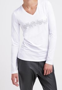 Bílé dlouhé rukávy tričko s výstřihem do V a šedým logem "ARMANIJEANS" přes přední část; hladký bavlněný materiál; upnutý střih; v kombinaci s černými kalhotami.