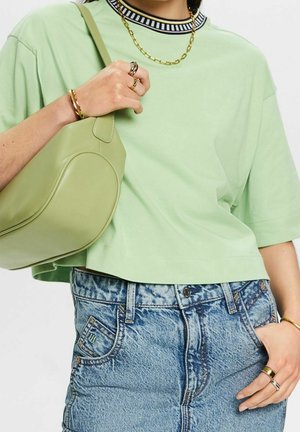 Person trägt ein hellgrünes T-Shirt, einen blauen Jeansrock, goldenen Schmuck und eine passende grüne Umhängetasche. Die Hand ruht in der Rocktasche.