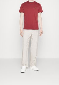 Rød bomulds T-shirt med rund hals og et lille gult logo, parret med lyse beige bukser og hvide sneakers.