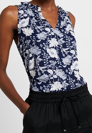 Sleeveless navy blue blouse with white floral print worn with black drawstring pants, model's hand in pocket.