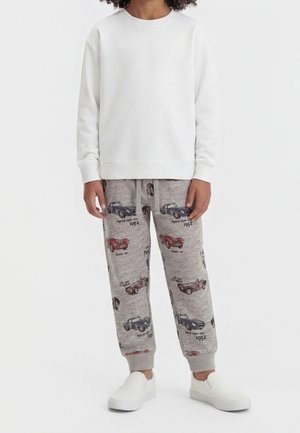 Barn iført en hvid langærmet skjorte, grå joggingbukser med vintage biltryk og hvide slip-on sko, står mod en hvid baggrund.