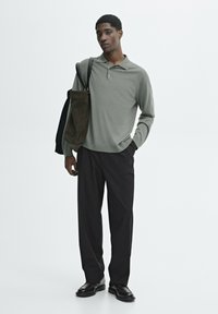 Long-sleeve, olive green polo shirt, black trousers, and black shoes. Model carries a dark brown suede tote bag over one shoulder.