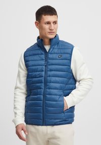 Mann trägt eine blaue gesteppten ärmellosen Weste über einem weißen Langarmshirt, hat eine Hand in der Westentasche und schaut zur Seite.