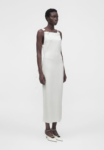 Γυναίκα που φοράει λευκό μίντι φόρεμα χωρίς μανίκια με floral λεπτομέρειες στα τιραντάκια, συνδυασμένο με λευκές μυτερές γόβες με λουράκια στον αστράγαλο.