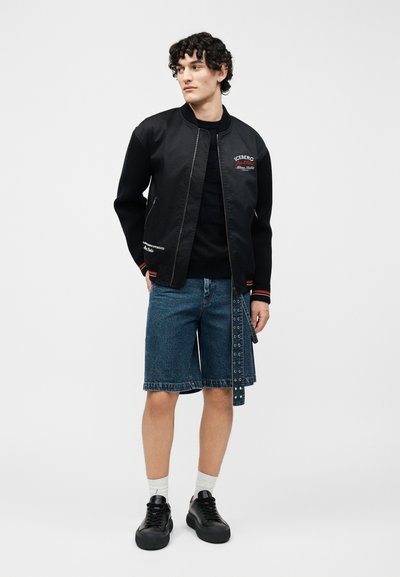 Νεαρός άνδρας με σκούρα σγουρά μαλλιά φοράει μαύρο μπουφάν με φερμουάρ, μαύρο πουκάμισο, μπλε τζιν σορτς με ζώνη με τρύπες, λευκές κάλτσες και μαύρα αθλητικά παπούτσια.