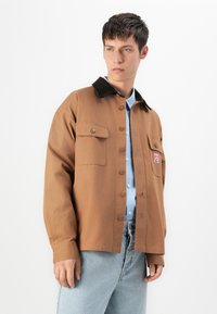 Joven vestido con una chaqueta marrón de botones con bolsillos en el pecho sobre una camisa de rayas azul y unos jeans de un azul claro, de pie contra un fondo blanco.