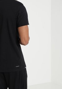 Svart t-shirt med kort ärm och rund halsringning, tillverkad av bomull, med en subtil logo på nedre rygg och en slät textur.