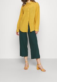 Camicia gialla con colletto, abbinata a pantaloni verdi a gamba larga con un motivo texturizzato e sandali gialli con tacco a blocco.