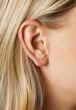 Blonde Haare hinter dem Ohr gesteckt mit kleinen runden Diamant-Steckerohrringen am Ohrläppchen auf heller Haut.