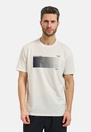 Uomo che indossa una T-shirt bianca a maniche corte Armani Exchange con linee orizzontali sfumate nere e pantaloni neri, in piedi davanti a uno sfondo semplice.