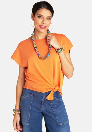 Woman wearing an orange short-sleeve top tied at the waist, blue jeans with large pockets, colorful beaded necklace, bracelets, and hoop earrings.