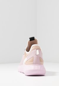 Chaussure de sport rose clair et beige vue de dos, montrant une semelle épaisse, des détails de couture et une languette de tirage noire sur le talon.