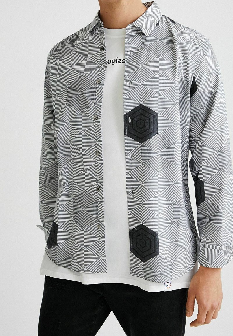 Černo-bílá košile na knoflíky se vzorem, který má geometrické tvary, a hexagonovými akcenty, s klasickým límcem. Nositá přes bílý tričko.