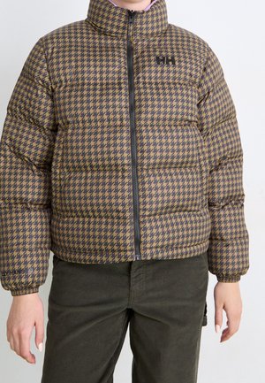 Persoon draagt een bruin-zwart geruit donzen jack met ritssluiting aan de voorkant, gecombineerd met donkergroene corduroy broek.