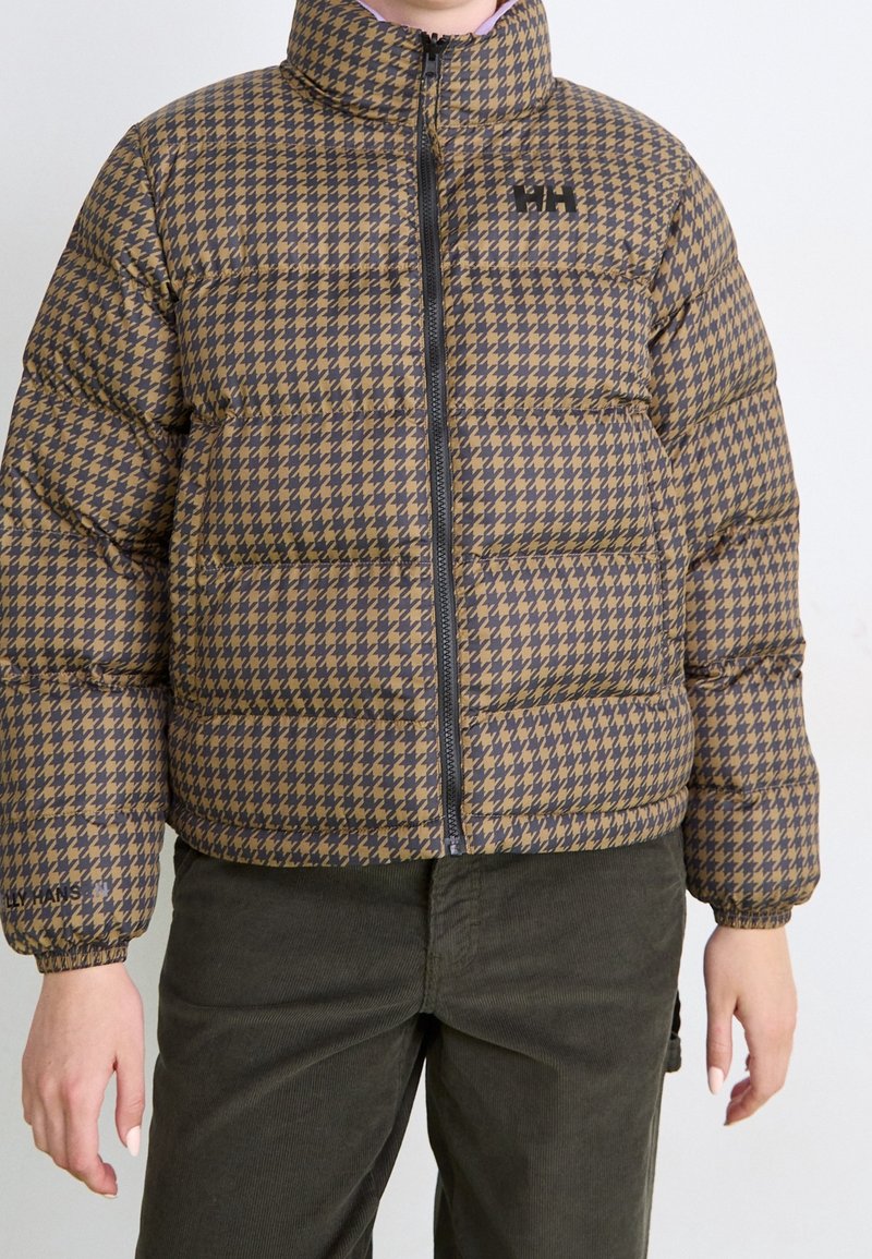 Personne portant une doudoune à motifs pied-de-poule marron et noir avec une fermeture éclair à l'avant, associée à un pantalon en velours côtelé vert foncé.