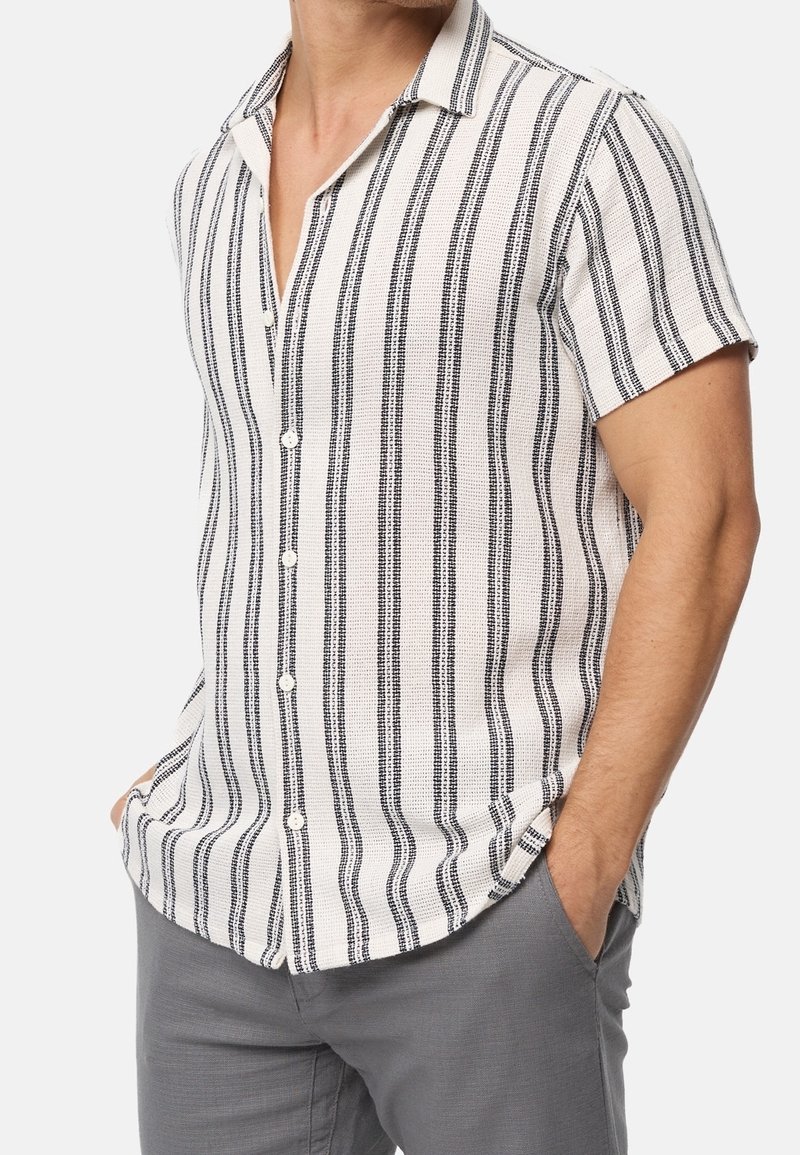 Homme portant une chemise blanche à manches courtes avec des rayures noires verticales et un pantalon gris, debout les mains dans les poches devant un fond uni.