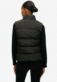 Chaleco acolchado negro con cuello alto, con secciones acolchadas horizontales y logo en relieve en el hombro izquierdo, combinado con ropa oscura.