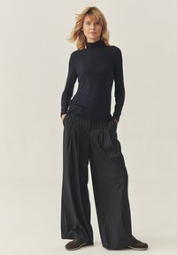 Mornarica majica dugih rukava s visokim vratom u kombinaciji s crnim hlačama širokih nogavica s prugama. Oba komada imaju opušten kroj i minimalne detalje.