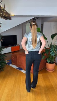 Λαμπερό ασημένιο κοντομάνικο τοπ με άνετη εφαρμογή, σε συνδυασμό με μαύρο τζιν με καμπάνα. Το δωμάτιο περιλαμβάνει τηλεόραση, γλάστρα με φυτό και διακοσμητικό καναπέ.