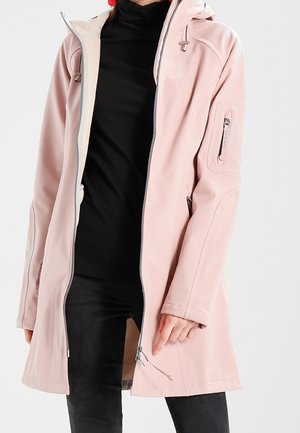 Osoba ubrana w jasno różowy płaszcz z kapturem, na czarnym golfie i czarnych spodniach, ręka spoczywająca w pobliżu suwaka płaszcza.