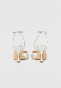Silver high-heeled sandals with ankle strap, open toe design, thin heel, and soft textured material. Beige sole with light detailing.