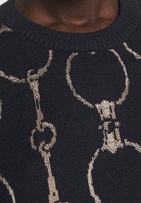 Námořnický pletený svetr se zlatým kovovým vzorem propojených řetězů, s žebrovaným kulatým výstřihem a texturovanou látkou.