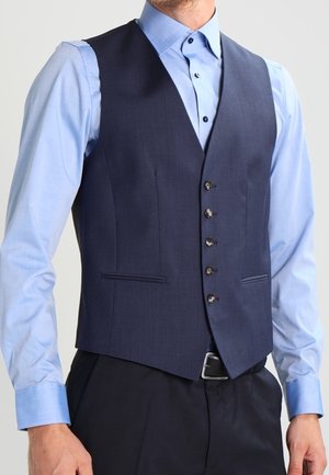 Mann trägt eine taillierte marineblaue Weste mit fünf Knöpfen über einem hellblauen Hemd und schwarzen Hosen, steht mit den Händen seitlich am Körper.