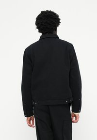 Chaqueta de denim negra con diseño de cuello, costuras visibles y cierres de botón a presión. Presenta un corte recto y costuras en los hombros.