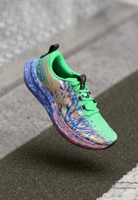 Leuchtend grüner Laufschuh mit blauer und orangefarbener Spritzdesign, blauer Sohle und schwarzem Ziehlaschen, im Flug über Asphalt festgehalten.