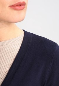 Granatowy kardigan w serek wykonany z miękkiego dzianiny, nałożony na jasnoszary sweter o prążkowanej fakturze.