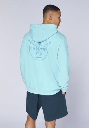 Mann mit dem Rücken zur Kamera, trägt einen hellblauen Hoodie mit Taucherlogo und dem Schriftzug „Chiemsee“, kombiniert mit dunklen Shorts, die Hände in den Taschen.