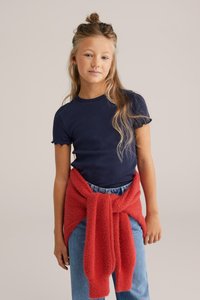 Námořnické pruhované tričko s krátkými volánkovými rukávy, doplněné zavázaným červeným svetrem. Světle modré džíny s klasickým střihem.