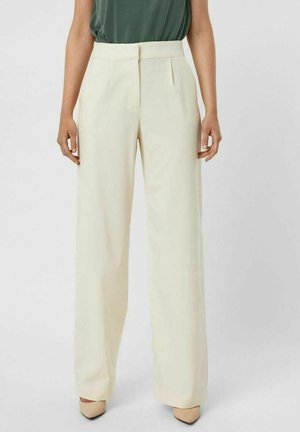 Femme portant un pantalon crème taille haute à jambes larges avec des plis, associé à des talons beige à bout pointu et un haut vert rentré.