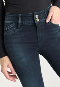 Hoge taille donker denim jeans met twee gouden knopen, één voorktas en een gladde textuur. De pasvorm is slank door het been.