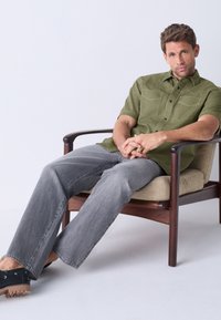 Green short-sleeve button-up shirt with dual chest pockets, worn with faded gray jeans and black shoes, seated on a wooden chair.