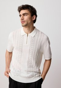 Polo de punto blanco con cuello con cremallera, detalles de patrón de hojas, mangas cortas y dobladillo acanalado. Textura suave.