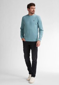 Jasnoniebieska bluza z okrągłym dekoltem i detalem w postaci logo, zestawiona z czarnymi dżinsami i białymi trampkami. Gładka tekstura, luźny krój.