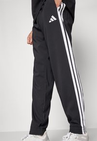 Fekete sportnadrág, három fehér csíkkal az oldalán, sima anyagból készült. Rugalmas mandzsettákkal rendelkezik, és Adidas logó található a csípőn.