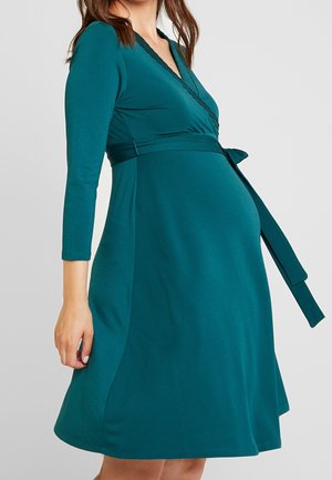 Schwangere Frau trägt ein tealfarbenes Wickelkleid mit dreiviertel Ärmeln und einem gebundenen Taillengürtel, steht vor einem hellen, schlichten Hintergrund.