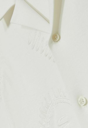 Camisa blanca con cierre de botón y un sutil bordado. La tela parece suave con una textura lisa y un diseño de cuello clásico.