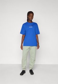 Blå bomullströja med korta ärmar och grafisk text "LONDON" tryckt framtill, kombinerad med ljusgröna sweatpants och svarta skor.