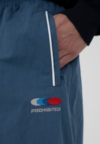 Main dans la poche d'un pantalon bleu marine avec un liseré blanc, montrant le logo brodé de trois ovales superposés en gris, bleu et rouge au-dessus du mot « INTERDIT ».