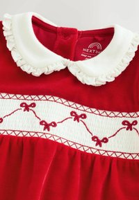 Abito rosso con colletto bianco a ruffles, fiocchi ricamati su un pannello bianco e tessuto testurizzato. Chiusura con bottoni sul retro.