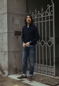 Tamsiai mėlynas užtraukiamas megztinis, balta palaidinė, mėlyni džinsai, rudi zomšiniai batai. Stovi prie akmeninės sienos ir puošnios metalinės vartų. Kasdieninė lauko aplinka.