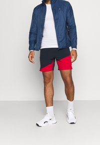 Chaqueta cortavientos marina, camiseta blanca, pantalones cortos a rayas en azul y rojo, zapatillas blancas y calcetines blancos. Conjunto de ropa deportiva exhibido.