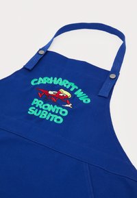Blue cotton apron with adjustable straps, featuring an embroidered cartoon design and text: “CARHARTT WIP PRONTO SUBITO” in green and red.