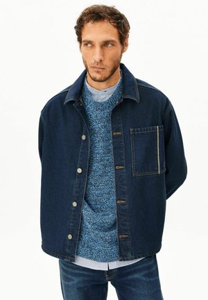 Mann mit kurzen lockigen Haaren, der eine dunkelblaue Jeansjacke über einem blau strukturierten Pullover und einem hellen Hemd trägt, vor einem einfarbigen weißen Hintergrund.