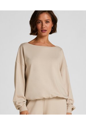 Donna che indossa una maglia beige larga a maniche lunghe con spalle scese e polsini a coste, in piedi contro uno sfondo chiaro e uniforme.