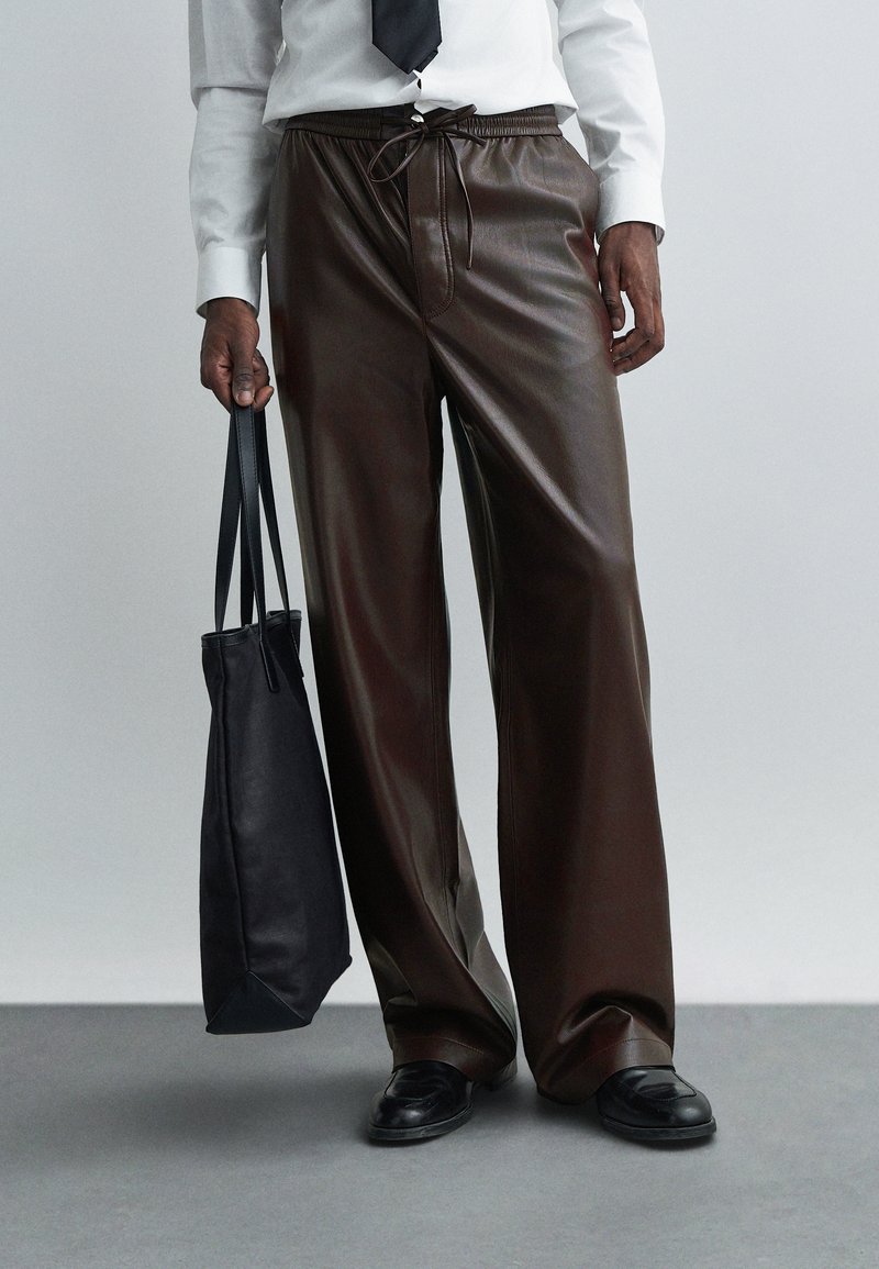 Oseba v temno rjavih usnjenih hlačah, beli majici, črni kravati in črnih čevljih, drži črno usnjeno torbo na sivoj ozadju.