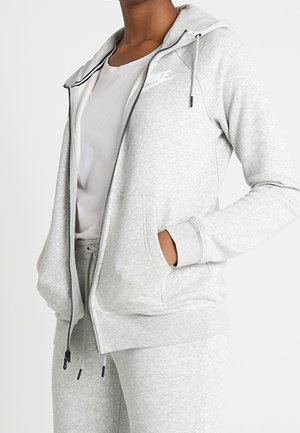 Personne portant un sweat à capuche zippé Nike gris clair avec les mains dans les poches et un pantalon de survêtement assorti par-dessus un t-shirt blanc.